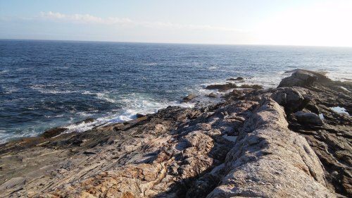 Rocky coastline with waves crashing against the shore under a clear blue sky. Sunlight glimmers on the water.