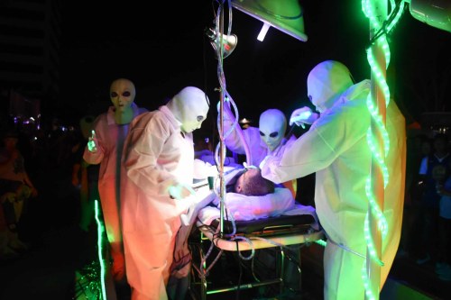 A group of aliens in white suits perform an operation on a person on a medical table, surrounded by colorful lights.