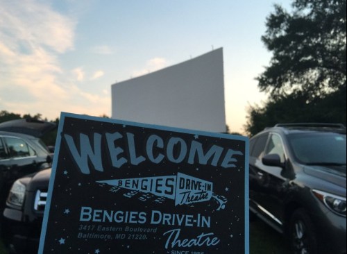 A welcome sign for Bengies Drive-In Theatre in Baltimore, with a blank movie screen and cars parked in the foreground.