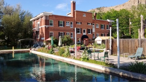 A brick building beside a clear pool, surrounded by greenery and outdoor seating in a sunny setting.