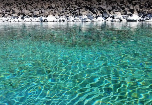 Clear turquoise water gently ripples over a rocky shoreline, reflecting light and creating a serene atmosphere.