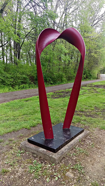 A large, abstract red sculpture resembling a heart shape, set on a black base in a green park area.