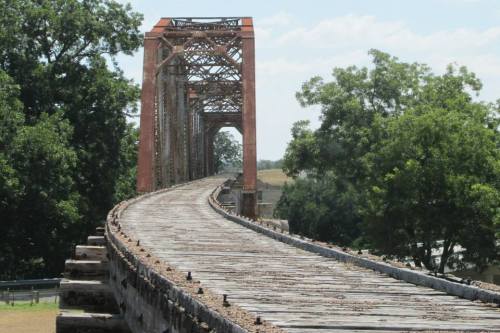 A weathered metal bridge stretches into the distance, surrounded by green trees and a clear blue sky.