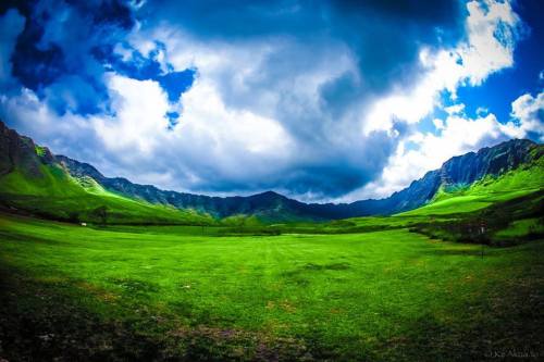 A panoramic view of a lush green valley surrounded by mountains under a dramatic sky with clouds.