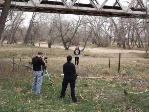 A person stands on a grassy area near a riverbank, while two others set up a camera on a tripod nearby.