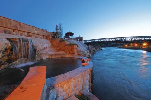 A serene riverside scene at dusk, featuring a waterfall, hot springs, and a bridge in the background.