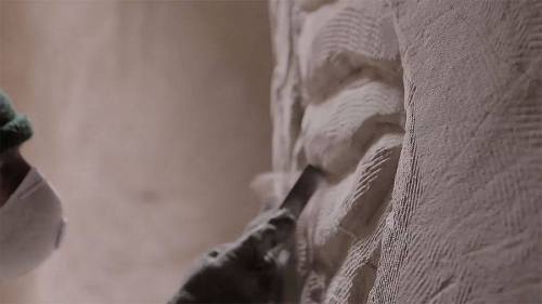 A person wearing a mask and gloves carves textured patterns into a wall with a tool.
