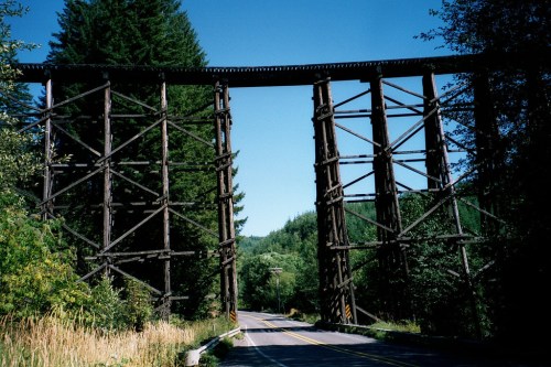 A tall wooden trestle bridge spans over a road, surrounded by lush green trees and a clear blue sky.