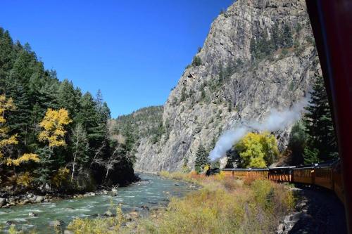 A scenic view of a train traveling along a river, surrounded by mountains and autumn-colored trees under a clear blue sky.