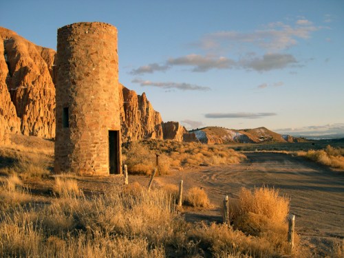 A stone tower stands beside a dirt road, surrounded by golden grass and rocky hills under a clear sky.