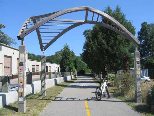 A bike path entrance marked by a curved metal arch, surrounded by trees and industrial buildings on a sunny day.