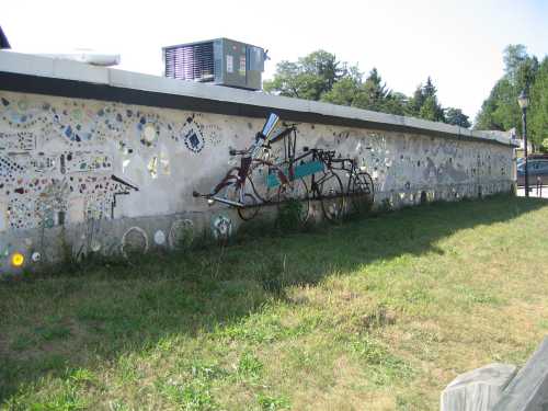 A colorful mural on a wall featuring various designs and a metal sculpture of a bicycle. Green grass in the foreground.