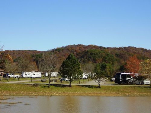 A serene campsite by a lake, featuring RVs, trees, and rolling hills with autumn foliage under a clear blue sky.