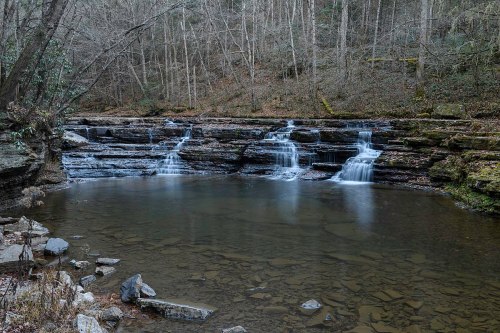 A serene waterfall cascades over rocky ledges into a calm pool, surrounded by trees in a tranquil forest setting.