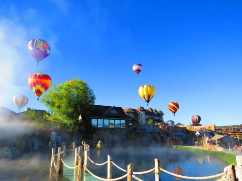 Colorful hot air balloons float over a serene landscape with a misty lake and a charming building in the background.