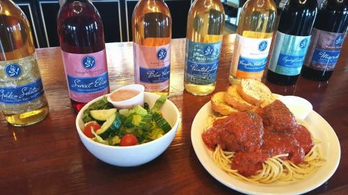 A plate of spaghetti and meatballs with a side salad, accompanied by several bottles of wine on a wooden table.
