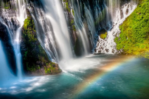A vibrant waterfall cascades into a turquoise pool, with a rainbow arching over the water amidst lush greenery.