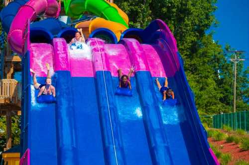 Three people slide down colorful water slides, splashing into a pool under a clear blue sky.