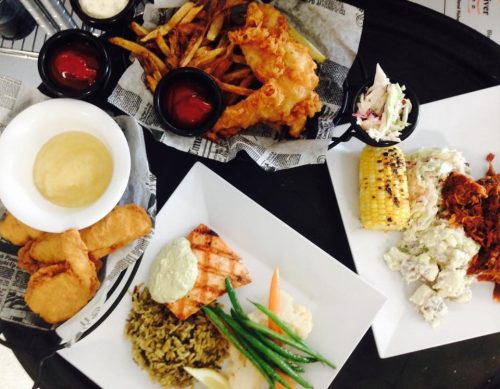 A variety of dishes including fried fish, fries, salmon, rice, coleslaw, corn, and a creamy dip on a black table.