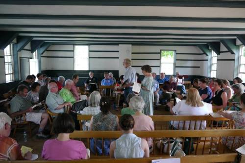 A group of people seated in a spacious room, engaged in a meeting or discussion, with a speaker at the front.