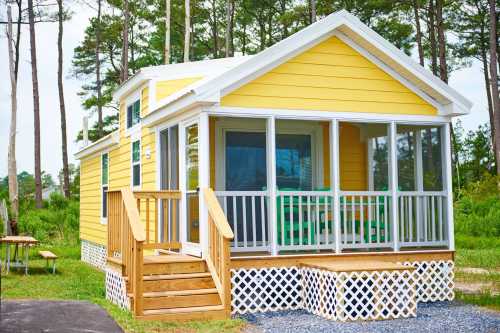 A bright yellow cottage with a porch, surrounded by trees and greenery, featuring a wooden ramp and white lattice skirting.