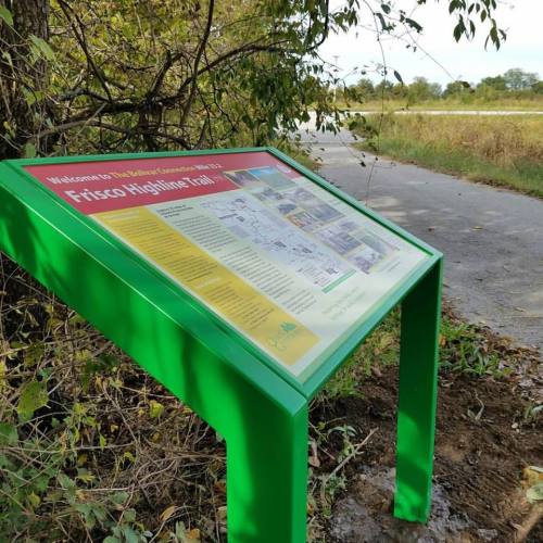 A green informational sign for the Frisco Highline Trail, surrounded by trees and grass, near a pathway.