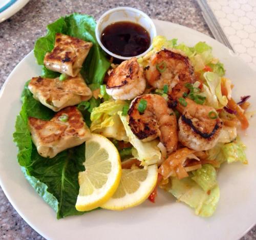 A plate of grilled shrimp on a bed of lettuce, accompanied by dumplings and a small bowl of dipping sauce.