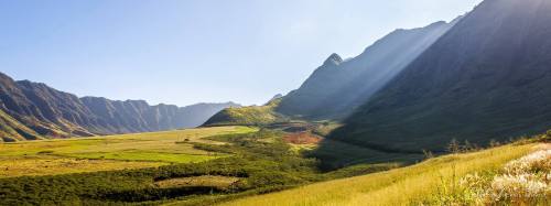 A panoramic view of lush green valleys and mountains under a clear blue sky, with sunlight streaming through.