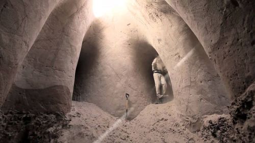 A dimly lit cave with a person standing on a mound of sand, holding a shovel in a sandy environment.