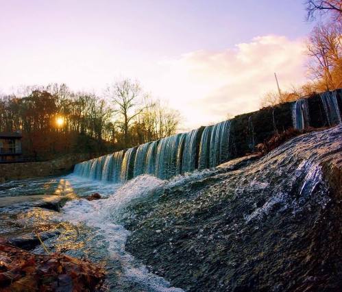 A serene waterfall cascades over rocks, with the sun setting behind trees in the background, creating a tranquil scene.