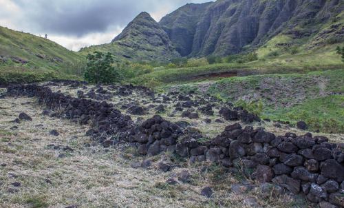 A rocky landscape with stone walls and lush green mountains under a cloudy sky.