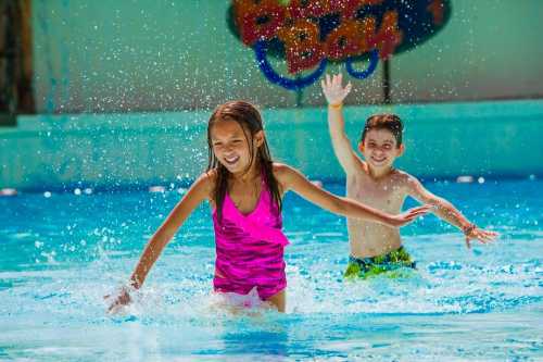 Two children play joyfully in a pool, splashing water and smiling under the sun.