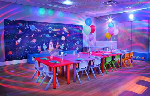 Colorful party room with a space-themed mural, decorated tables, balloons, and vibrant lighting effects.