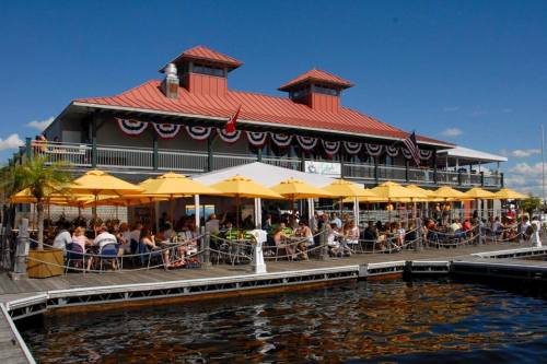A waterfront restaurant with a red-roofed building, outdoor seating under yellow umbrellas, and festive decorations.