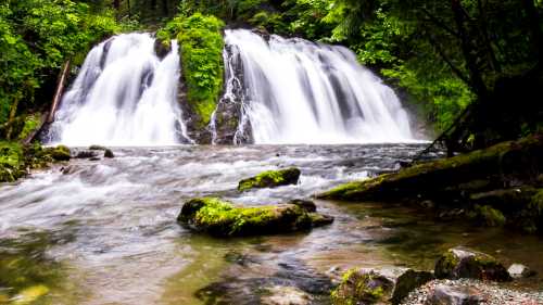 A serene waterfall cascades over rocks, surrounded by lush green foliage and a gentle river flowing below.