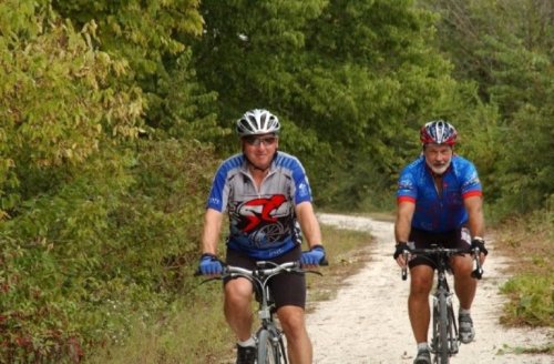Two men riding bicycles on a dirt path surrounded by greenery. Both are wearing helmets and cycling jerseys.