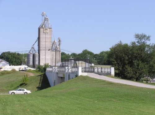 Grain silos stand tall against a clear blue sky, with a grassy slope and a white car parked nearby.