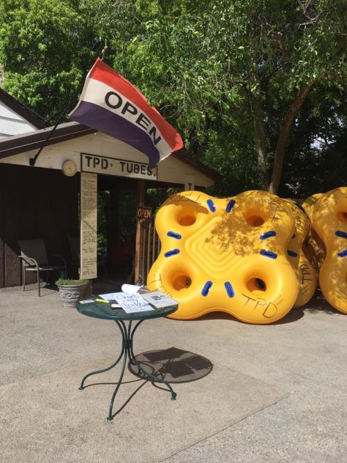 A rental shop with a flag reading "OPEN" and large yellow inner tubes outside, surrounded by greenery.