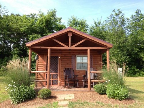 A cozy wooden cabin with a porch, surrounded by greenery and decorative plants.
