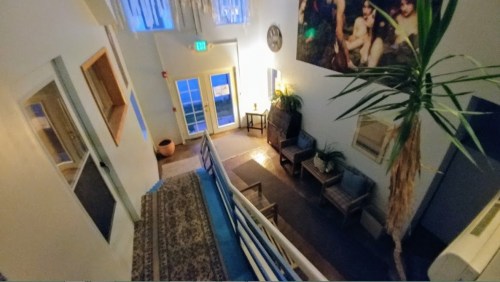 A cozy hallway with a staircase, plants, and seating, illuminated by natural light from large windows.
