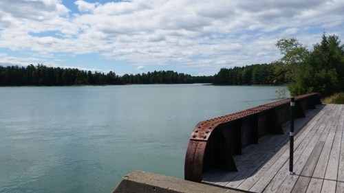 A calm lake surrounded by trees under a cloudy sky, with a wooden dock featuring a rusted metal railing.
