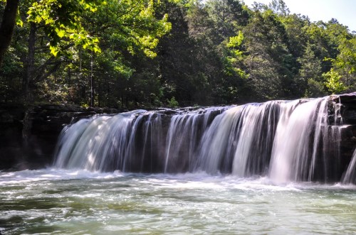 A serene waterfall cascades over rocks into a calm, clear pool, surrounded by lush green trees.