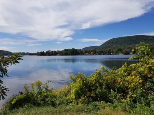 A serene lake surrounded by greenery and hills under a blue sky with fluffy clouds.