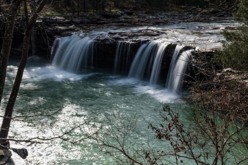 A serene waterfall cascades over rocks into a tranquil turquoise pool, surrounded by trees and natural scenery.