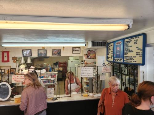 A busy diner with a counter, menu board, and customers ordering food. A staff member is serving behind the counter.