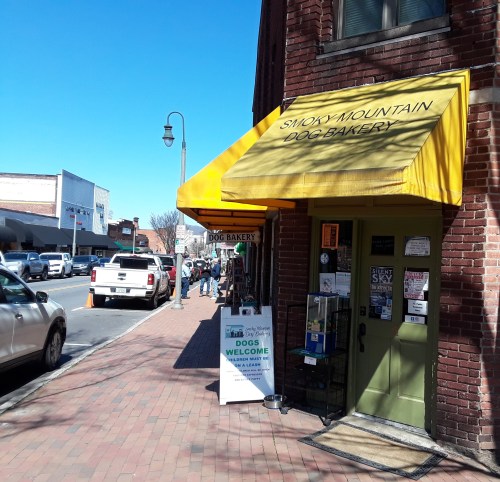 A yellow awning marks the entrance of Smoky Mountain Dog Bakery on a sunny street with pedestrians and parked cars.