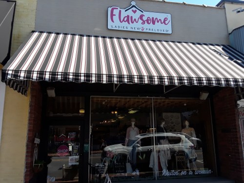 Exterior of a boutique named "Flawsome," featuring a striped awning and a display of women's clothing in the window.