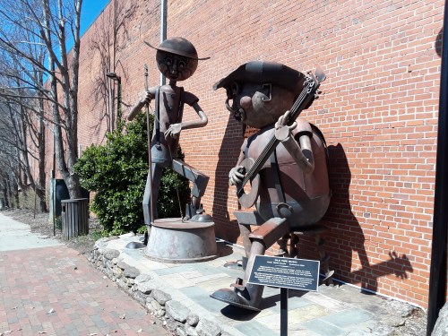 Two whimsical metal sculptures of musicians playing instruments, set against a brick wall with greenery nearby.
