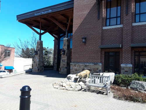 A stone building entrance with wooden beams, a bench, and a sculpture of a dog on a landscaped path.