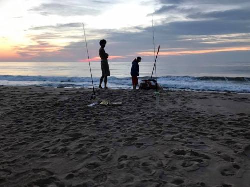 Two people fishing on a beach at sunset, with fishing rods and gentle waves in the background.